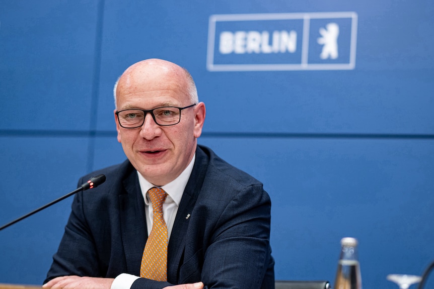 A balding man in a suit sits behind a microphone.
