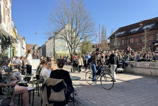 A busy street in Aarhus, with people sitting outside cafes, a cyclist riding down a path, and people gathered in a public space.