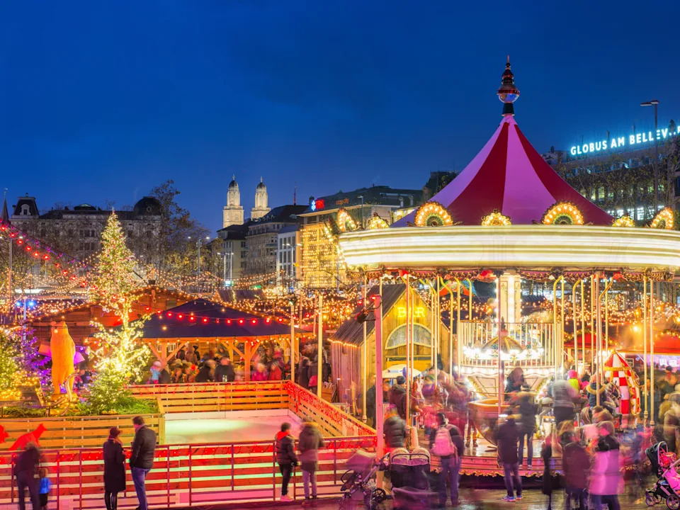 Festive Christmas market with illuminated carousel, trees, and crowds in a city square at dusk.