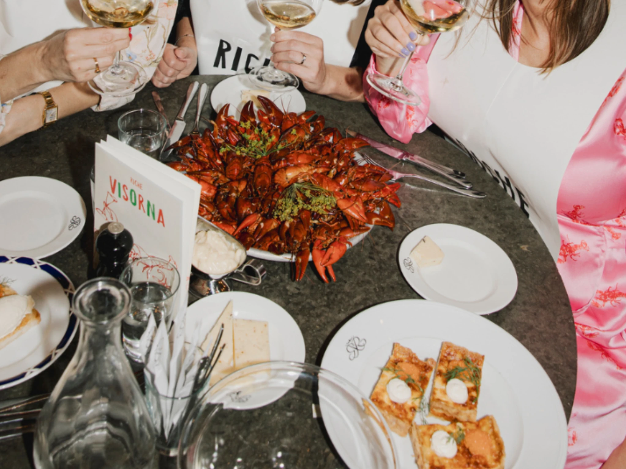 People wearing paper bibs clinking wine glasses over a table filled with red crayfish, Västerbotten cheese pie, and a songbook at Restaurant Riche.