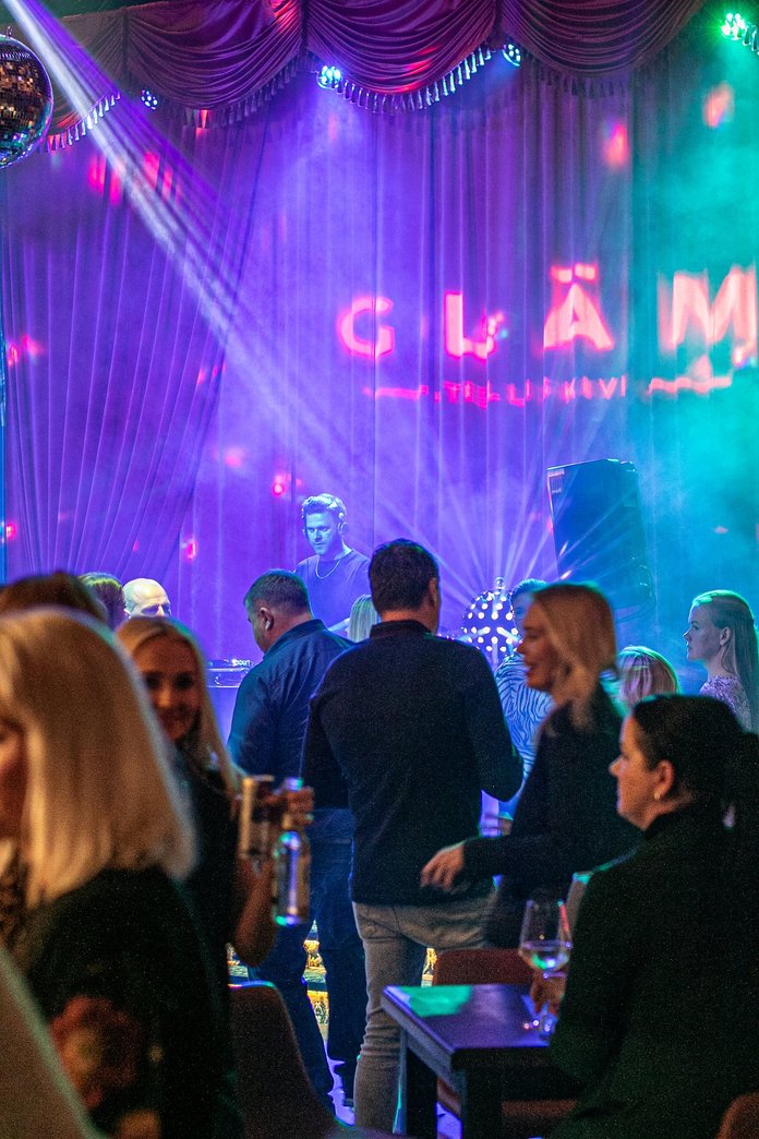 Üritus KeskeaBriis lokaalis Gläm. Jürgen on DJ-puldis, Grete kõnnib läbi saali.