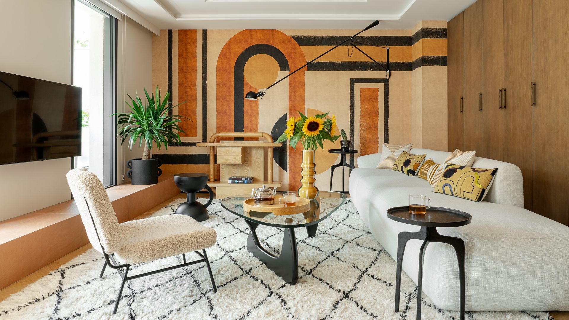 Salón con sofá blanco, pared con mural de papel pintado en colores tierra, alfombra, mesa de centro de diseño con el sobre de cristal, jarrón con girasoles,  butaca,  televisión colgada en la pared, techos con molduras