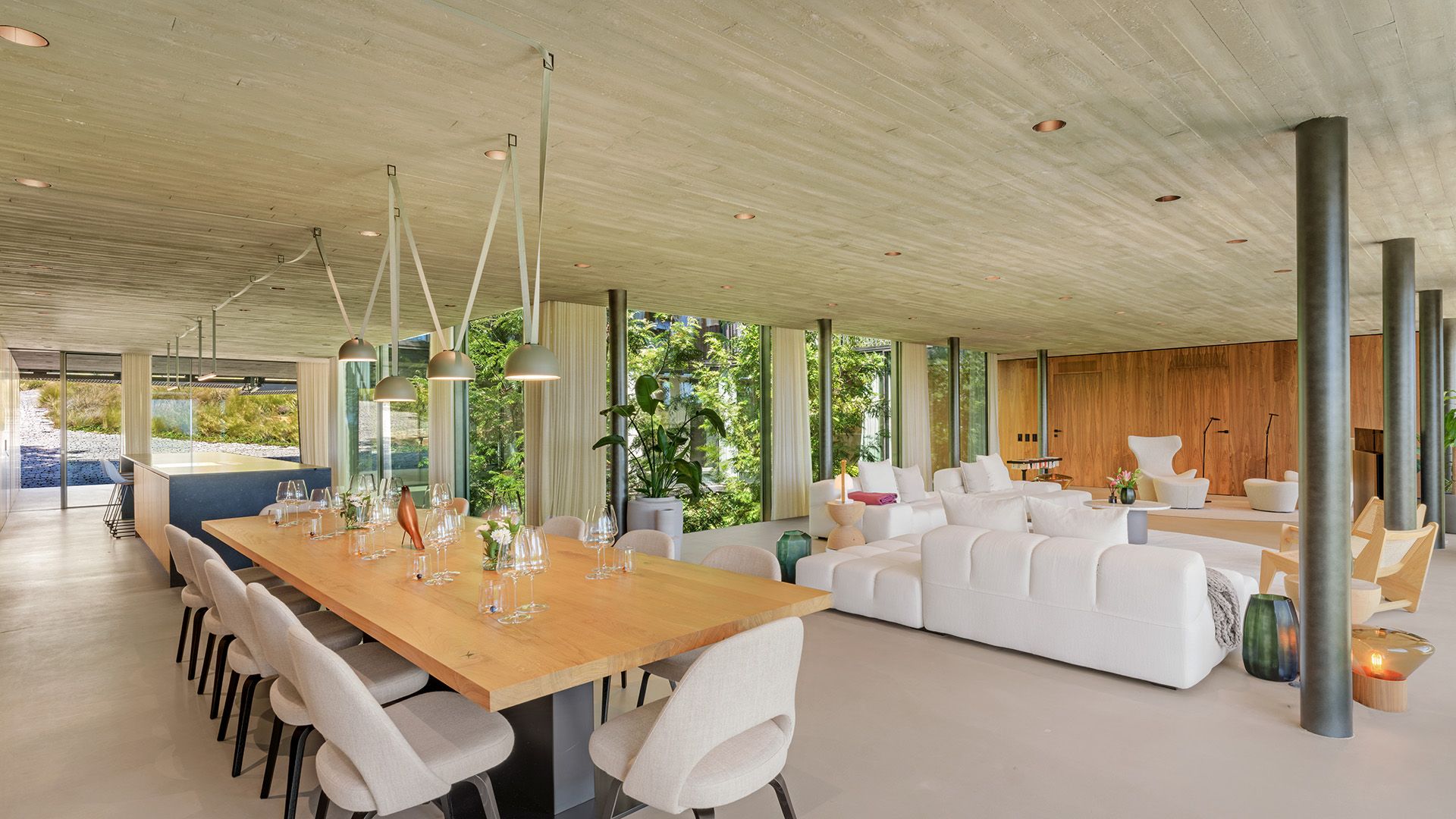 Espacio único que alberga salón, comedor y cocina, con techo de hormigón, pared de madera, columnas y tabiques de cristal 