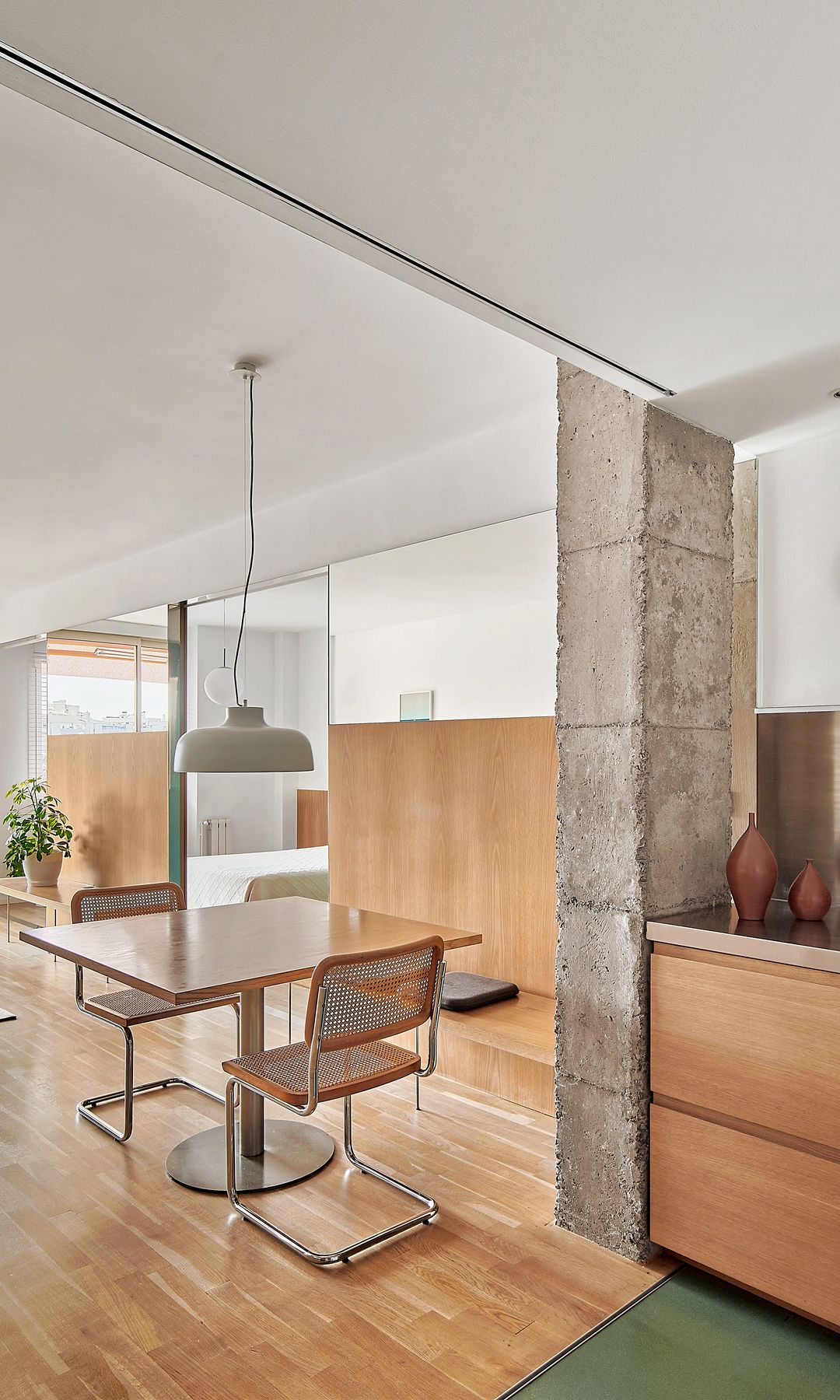Cocina abierta al comedor y al salón, con suelo de madera, banco, mesa de madera, sillas Cesca, lámpara de techo