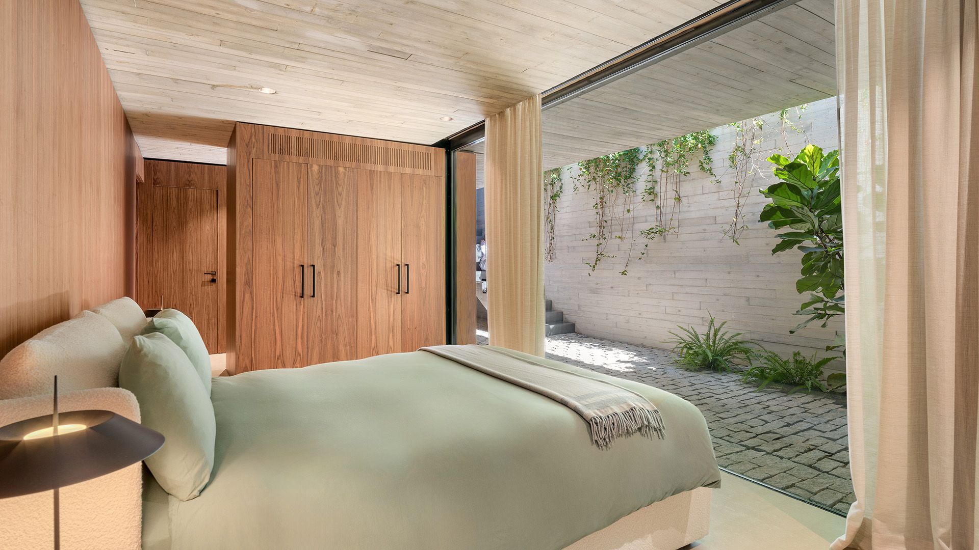 Dormitorio con salida al jardín, gran ventanal y cortinas beiges, armario de madera, ropa de cama verde oliva, techo de hormigón 
