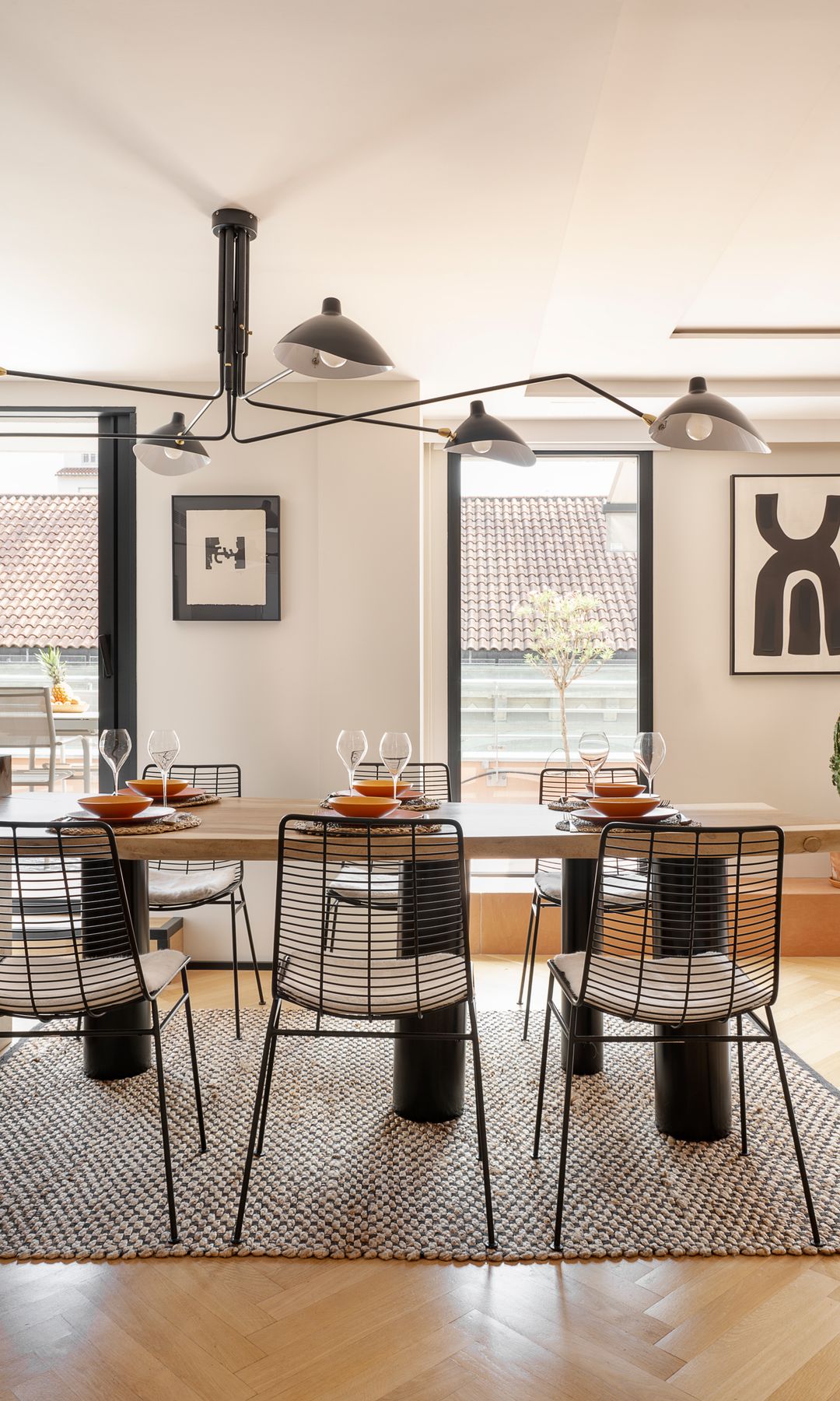 Comedor con mesa de madera y patas negras, alfombra de fibras vegetales, lámpara de techo, suelo de madera, cuadros en la pared, mueble bajo de madera, 