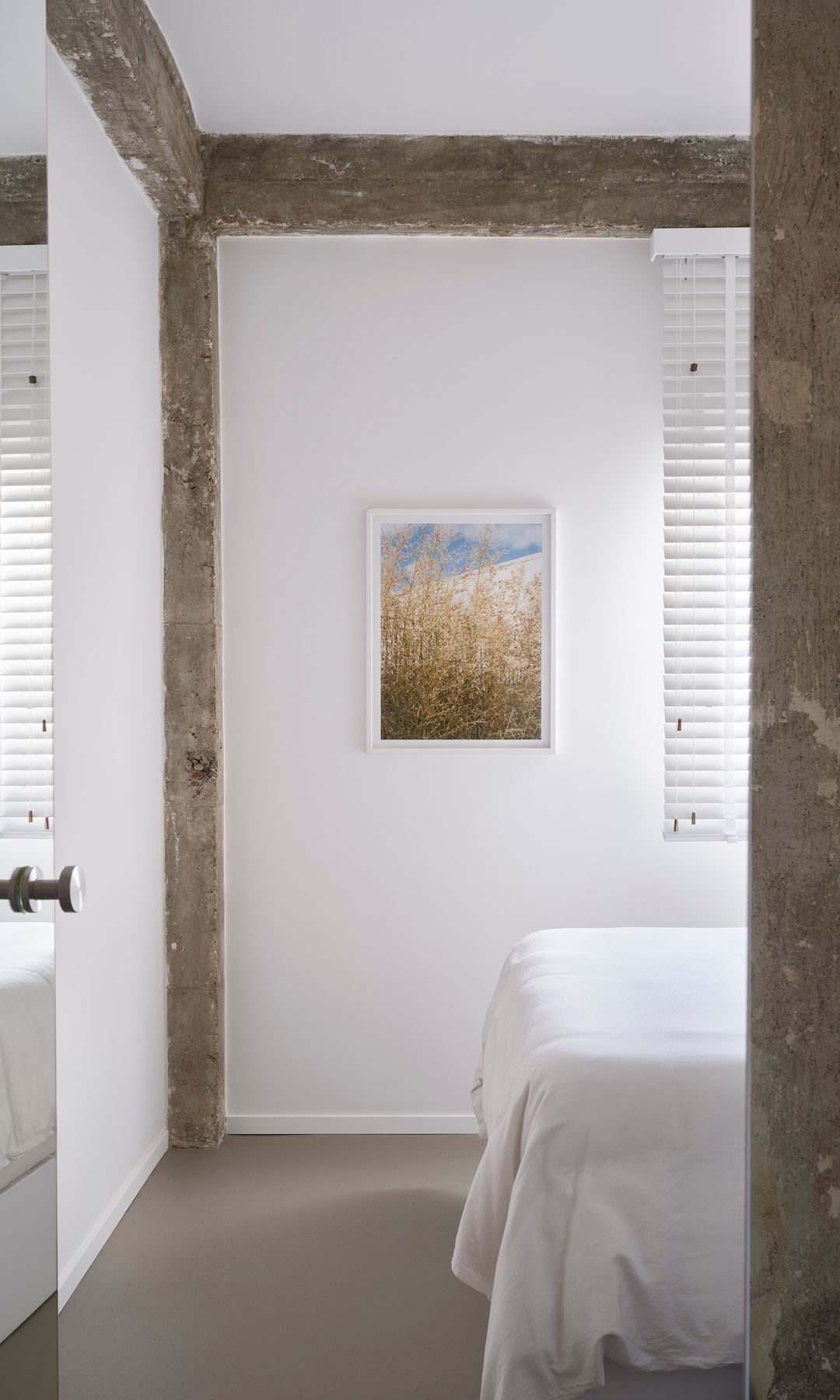 Dormitorio con columnas y vigas de hormigón, cuadro en la pared, estor blanco y ropa de cama blanca
