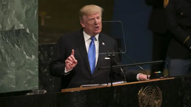 Donald Trump habla ante la Asamblea General de la ONU.  Viste un traje oscura, camisa blanca y corbata celeste. Apoya la mano izquierda sobre el podio y tiene la derecha levantada con el dedo índice señalando hacia arriba.