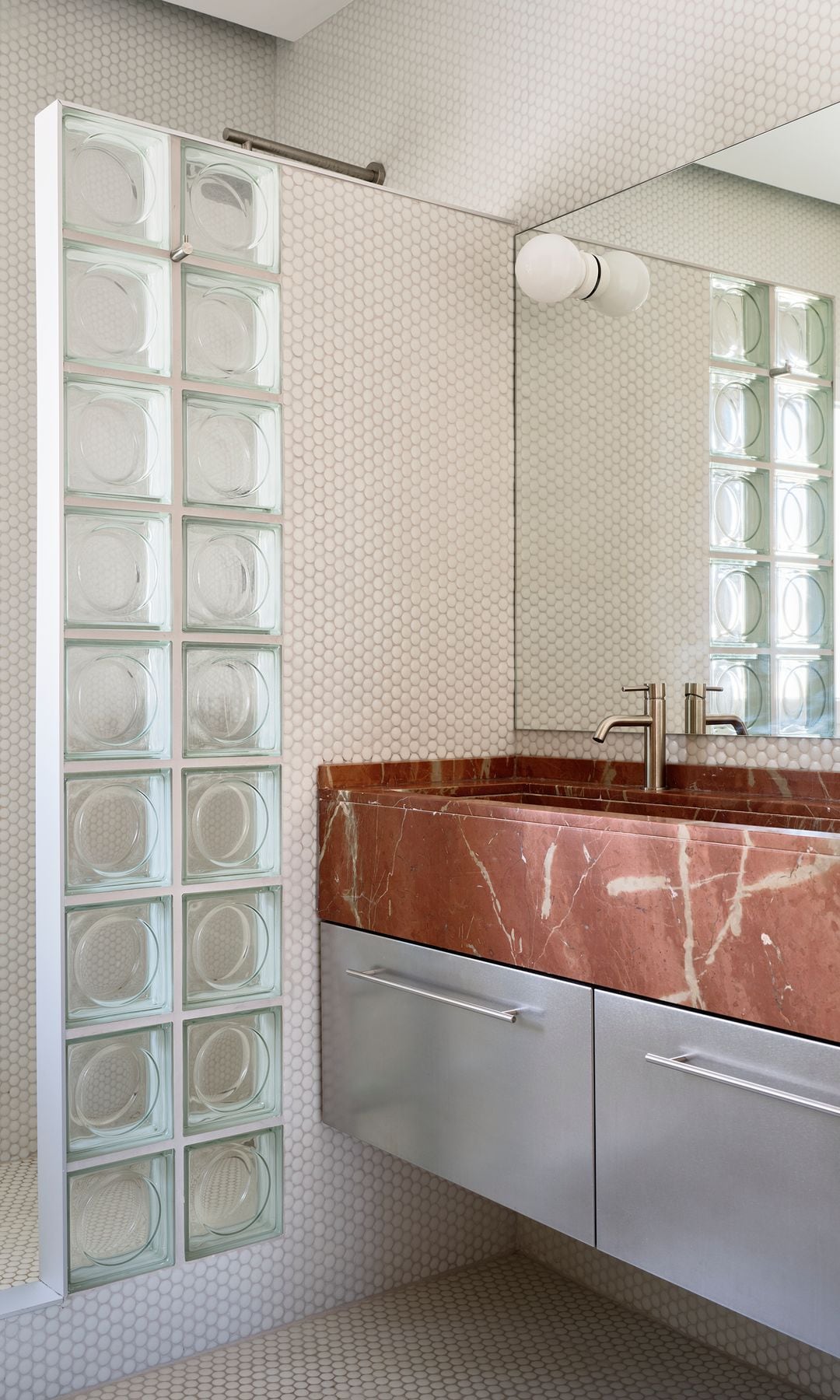 Baño con encimera de piedra roja, espejo, mueble metálico, ducha tras pared de pavés