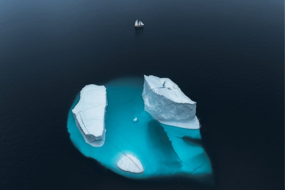 Imagen aérea de dos icebergs en Groenlandia, captada por Thorarinn Jonsson. La parte sumergida, de color turquesa, destaca gracias al ángulo de la toma y la intensidad de la luz polar. Un velero pequeño aparece en la distancia, usando gran campo de visión para comparar tamaños.