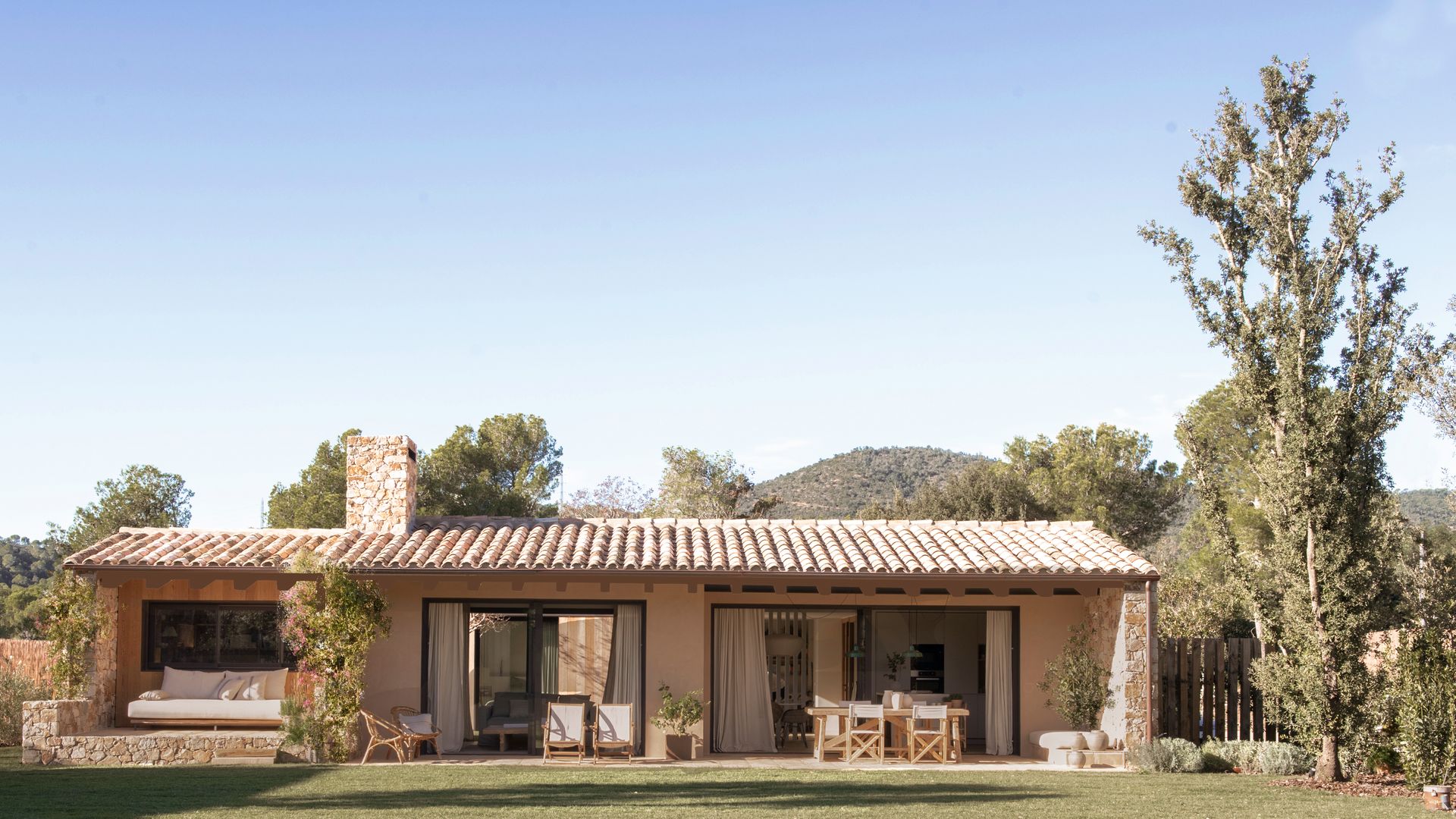 Fachada casa unifamiliar en una sola planta, con tabiques de cristal, jardín, muebles de madera, árboles y zona de estar, comedor y relax