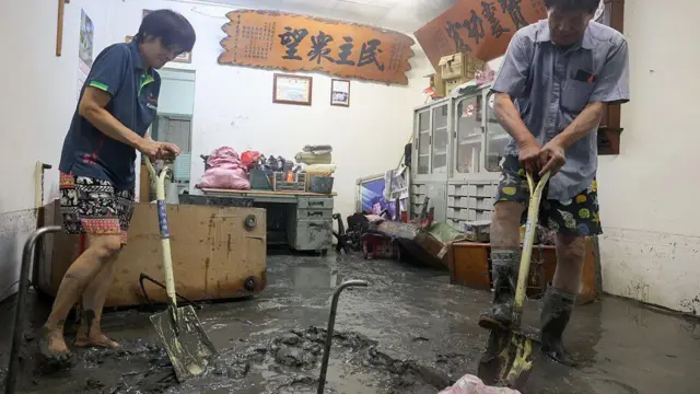 Dos personas limpian con palas el barro que se acumula en un local y que ha dañado mobiliario y electrodomésticos.