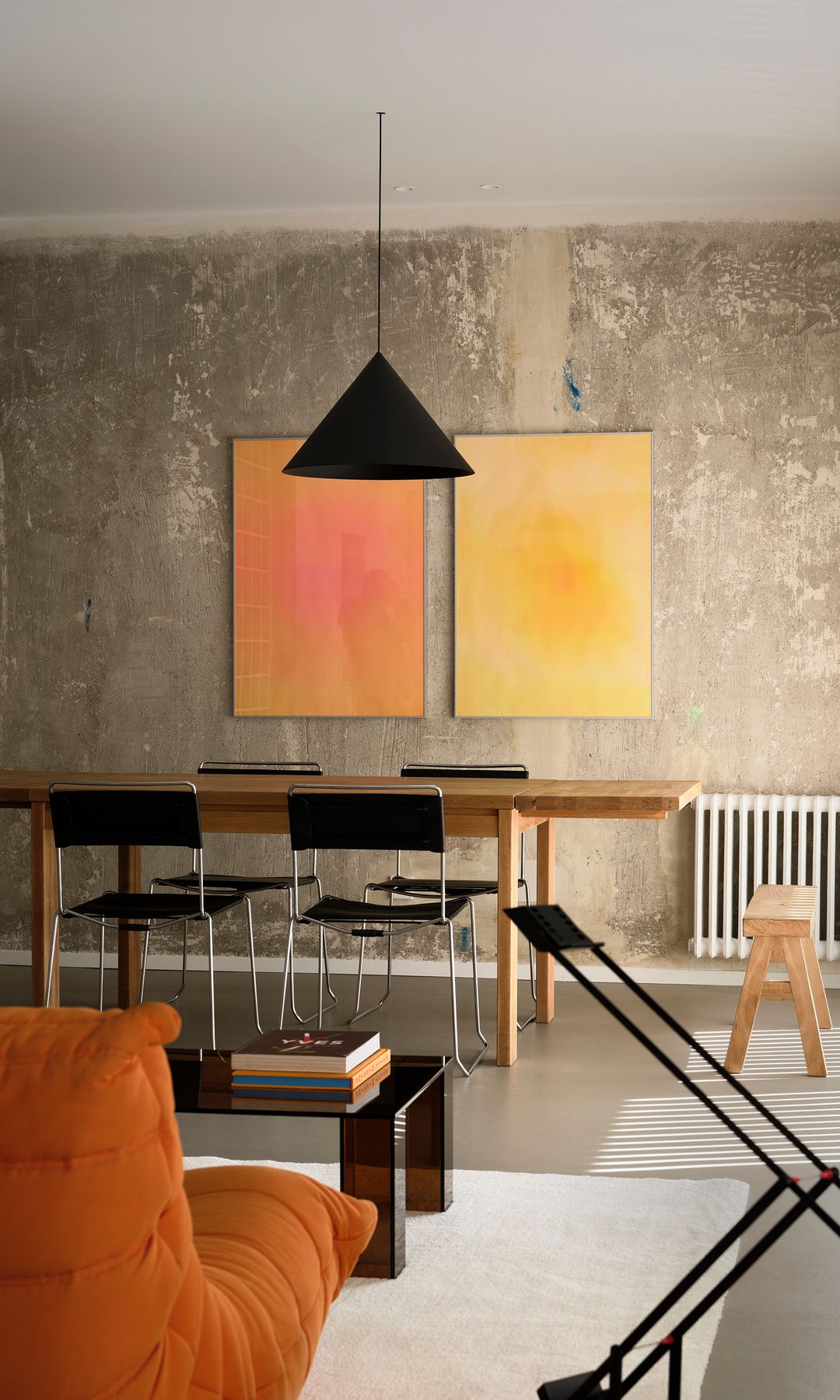 Salón comedor con mesa de comedor de madera, pared de hormigón, lámpara de techo negra, cuadros en la pared, sillas metálicas y negras, taburete de madera
