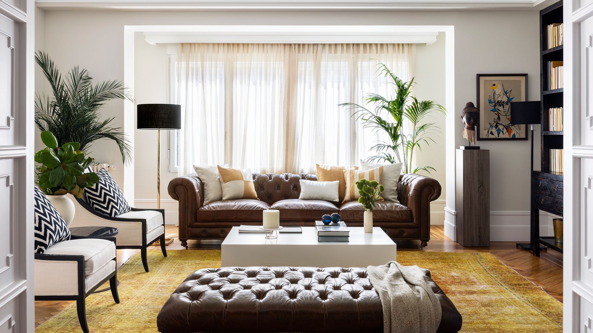 Salón con sofá Chester de cuero en marrón oscuro, banqueta de cuero a juego, alfombra, cortinas blancas, lámpara de pie, butacas tapizadas en blanco con cojín blanco y negro