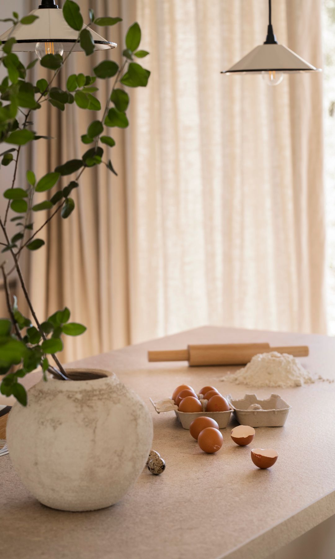 Isla de cocina con encimera blanca, lámpara de techo blanca, cartón de huevos, harina, rodillo y maceta con planta verde 