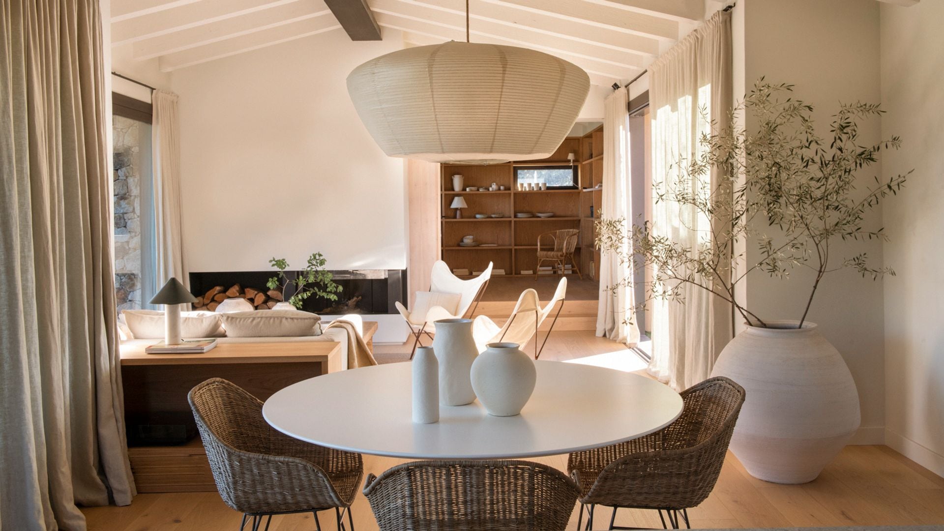 Salón comedor de estilo mediterráneo con vigas de madera, sillas de fibras vegetales, lámpara de techo de papel, suelo de madera, maceta grande blanca con olivo 