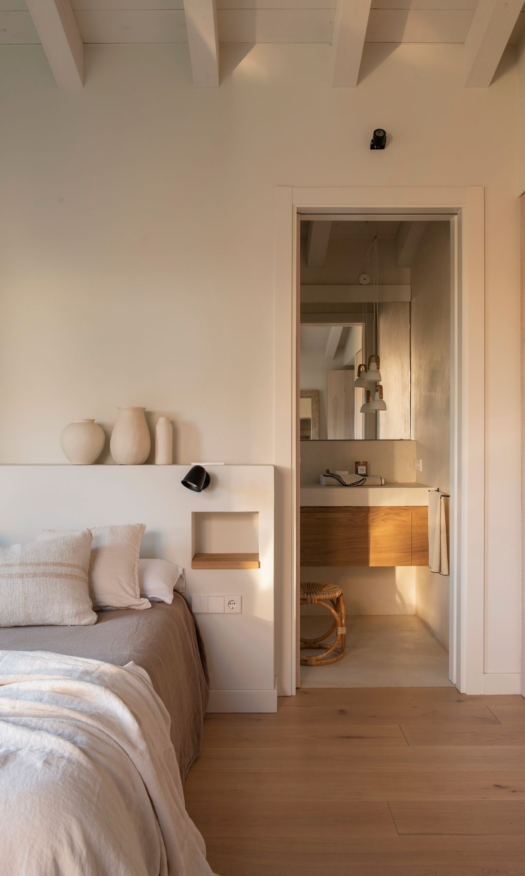 Dormitorio de estilo mediterráneo con baño, cabecero de obra con repisa, vigas en el techo, suelo de madera, aplique de pared negro