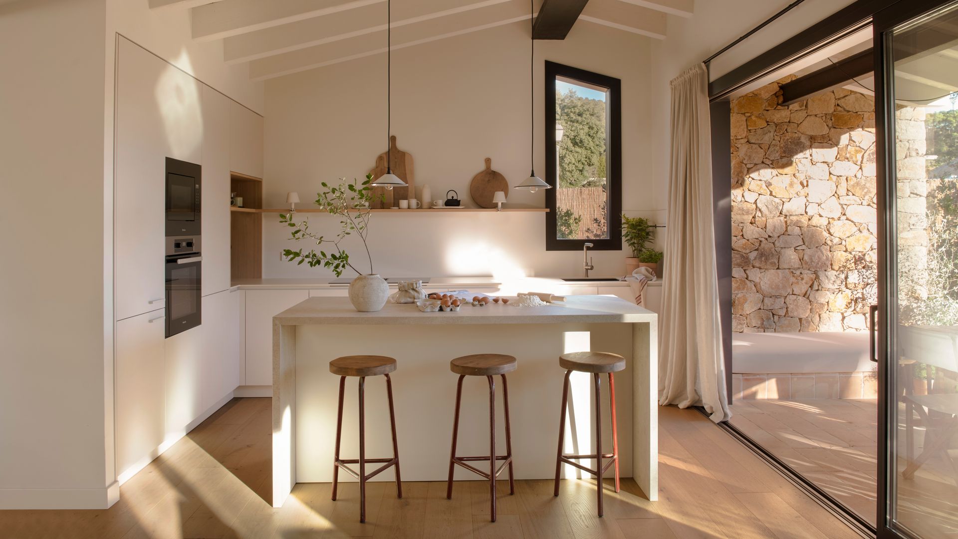 Cocina blanca con isla, con salida al jardín, taburetes de madera, vigas de madera blancas, suelo de madera, tablas y accesorios en una balda, cortinas 