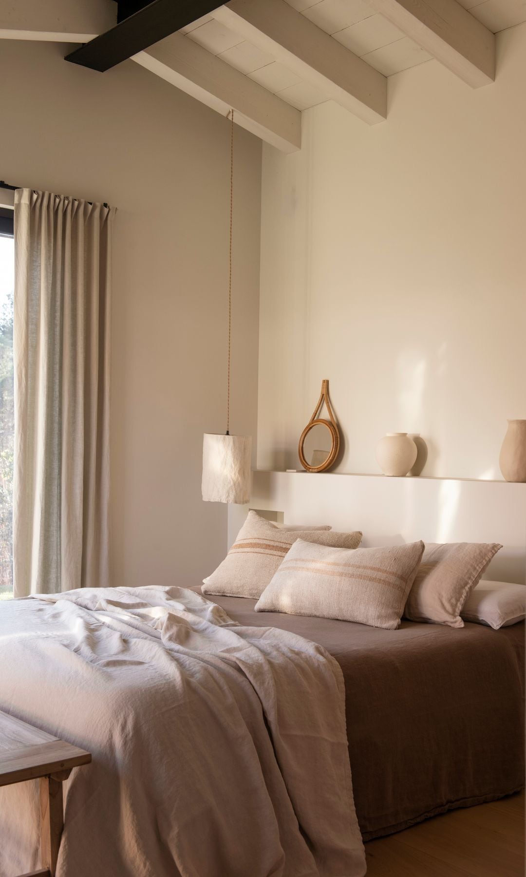Dormitorio con vigas de madera, cortinas cabecero de obra con repisa, ropa de cama marrón y beige, cojines, banco de madera a los pies de la cama