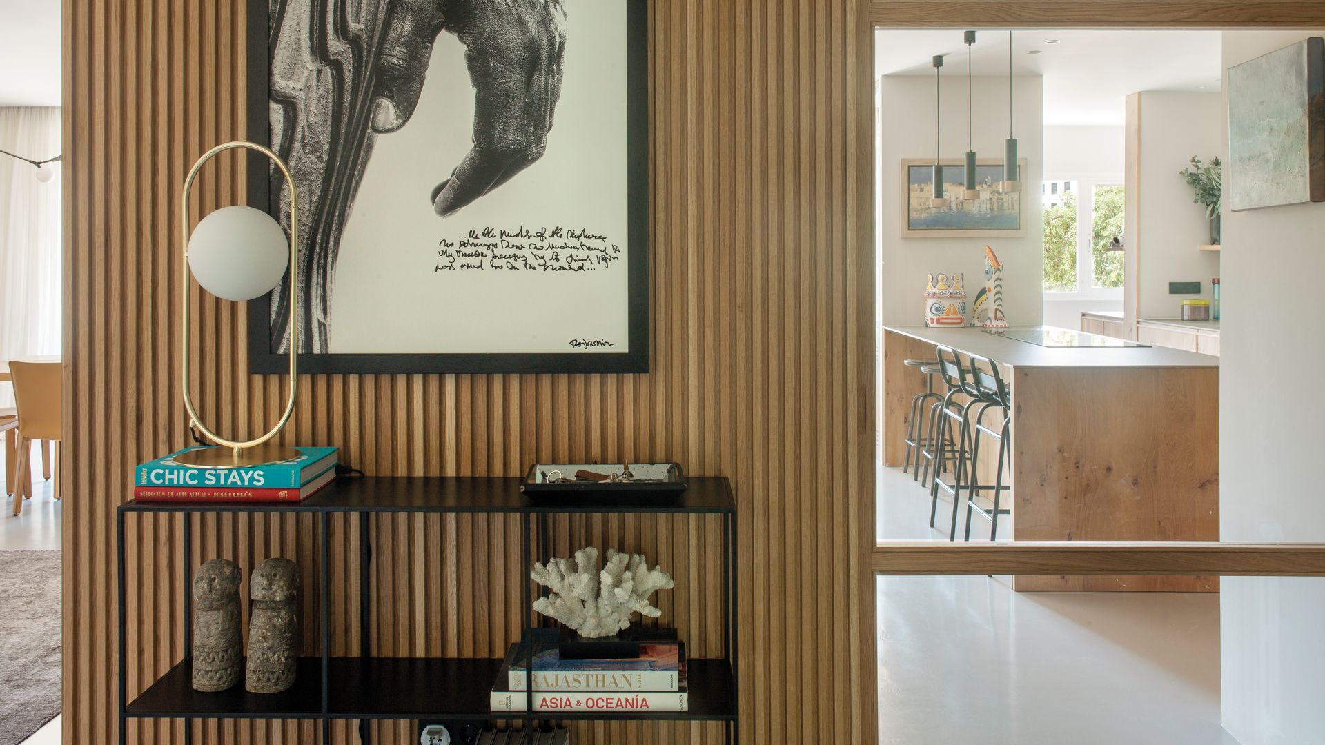 Recibidor con pared alistonada, puerta de cristal hacia la cocina, cuadro, consola metálica negra con libros, lámpara de sobremesa dorada y libros