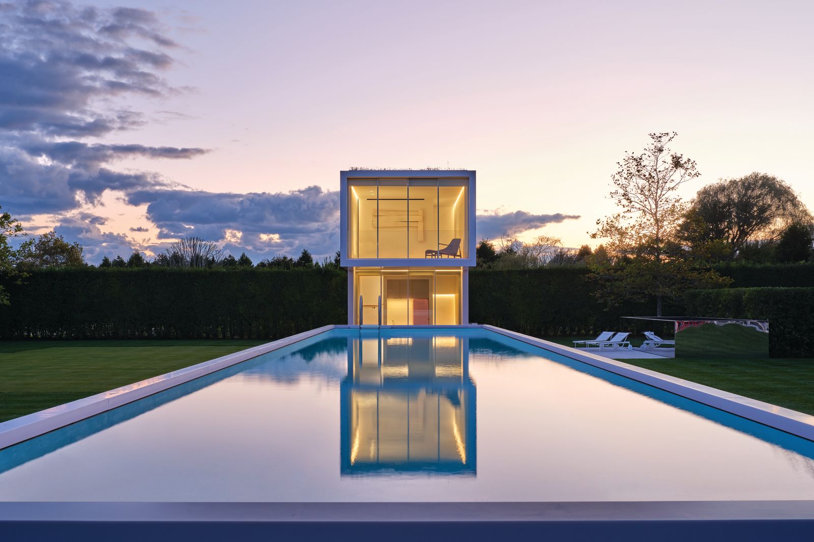 Casa minimalista con alberca gigante en el pasto refleja un atardecer