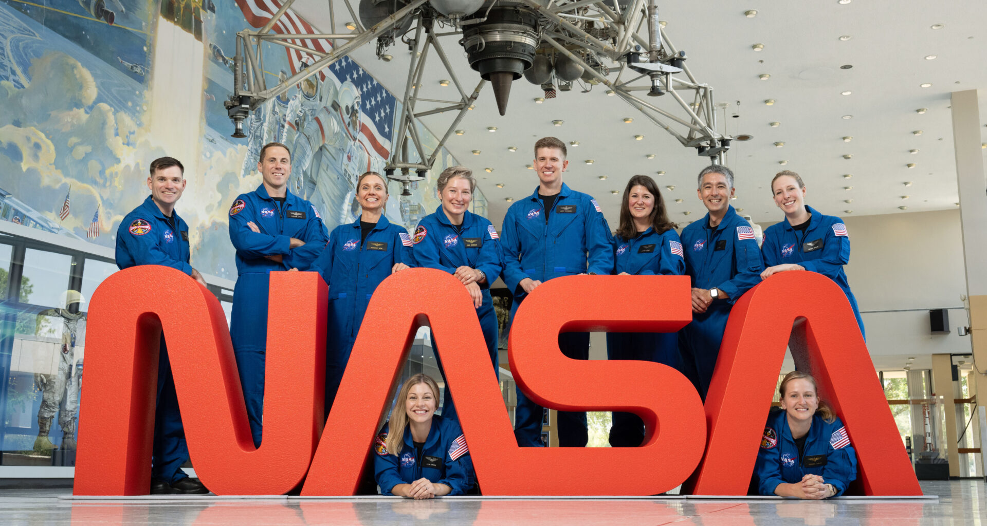 Ten members of NASA’s astronaut class in blue flight suits look and smile at the camera while gathered around a large NASA Worm logo.