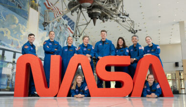 Ten members of NASA’s astronaut class in blue flight suits look and smile at the camera while gathered around a large NASA Worm logo.