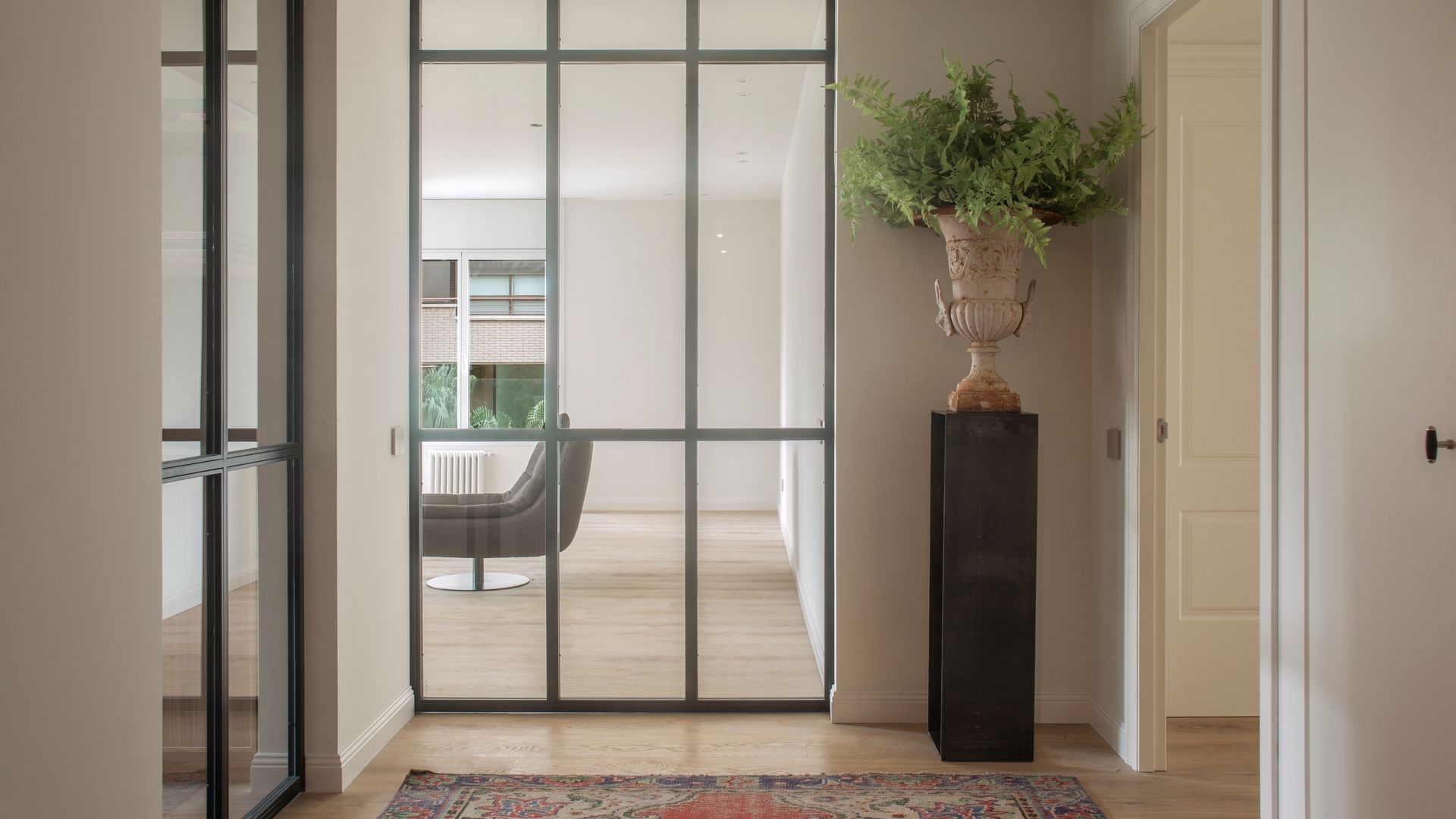 Recibidor con paredes de cristal con cuarterones negros, alfombra, pedestal con jarrón con helecho