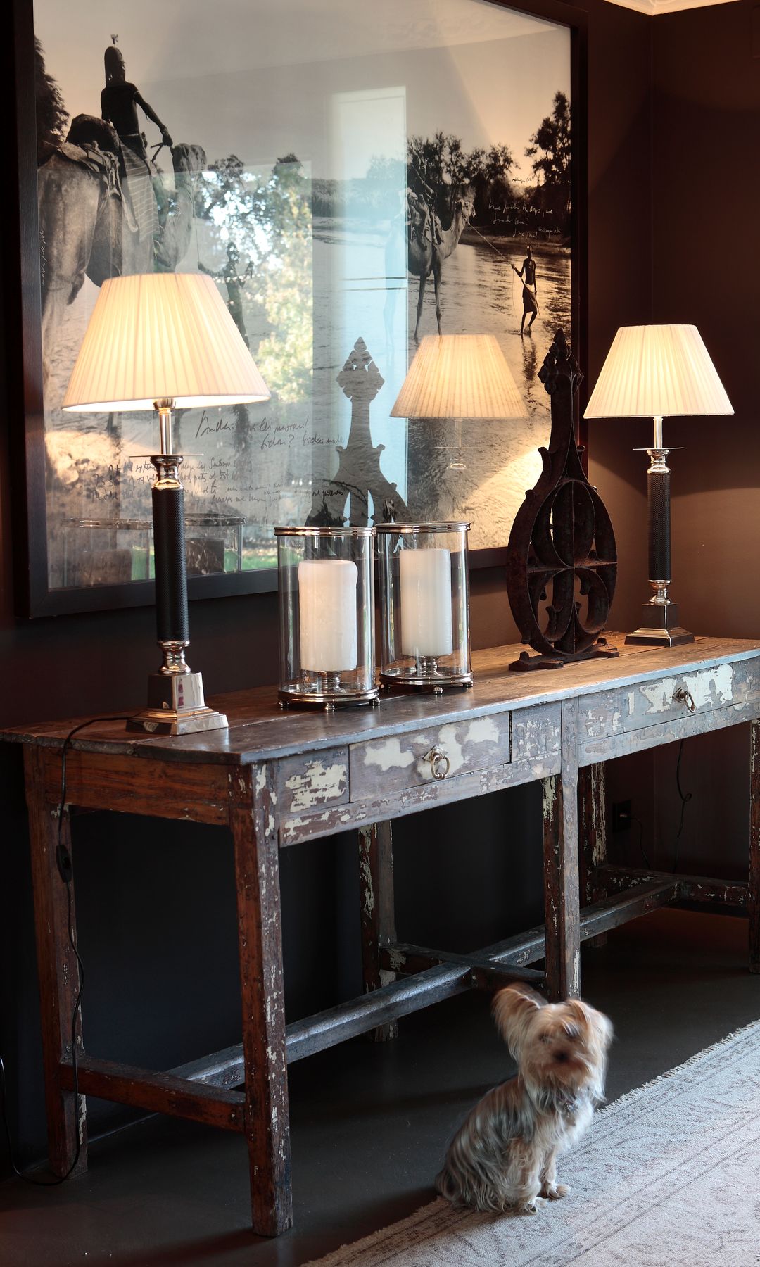 Recibidor con consola de madera antigua, lámparas de sobremesa y velas, fotografía XL en blanco y negro en la pared y perro pequeño en el suelo