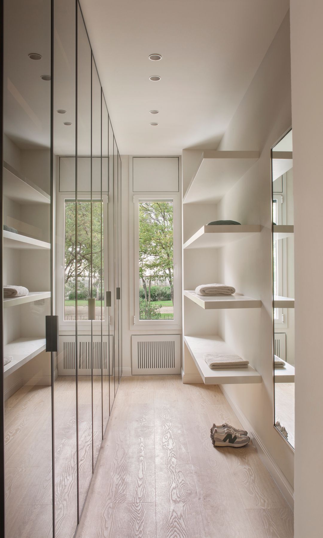 Vestidor con pared de armarios con puertas de cristal, suelo de madera, baldas blancas y ventana vertical