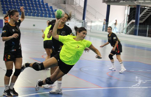 Una jugadora de balonmano en acción en un partido femenino en una pista cubierta, con otras jugadoras en el fondo y espectadores en las gradas.