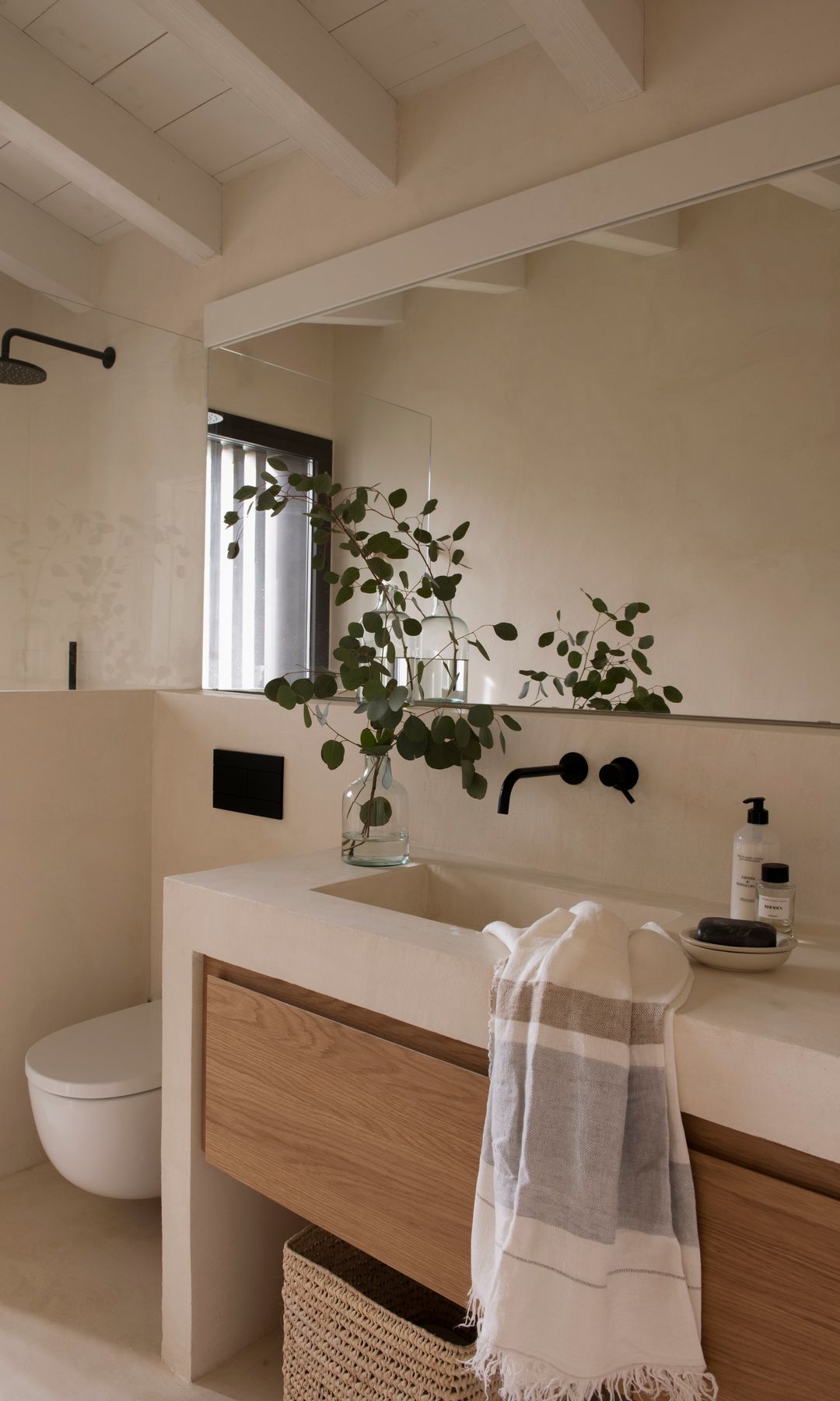 baño en blanco con mueble de madera, encimera blanca, jarrón de cristal con ramas, sanitario suspendido, espejo, grifería negra 