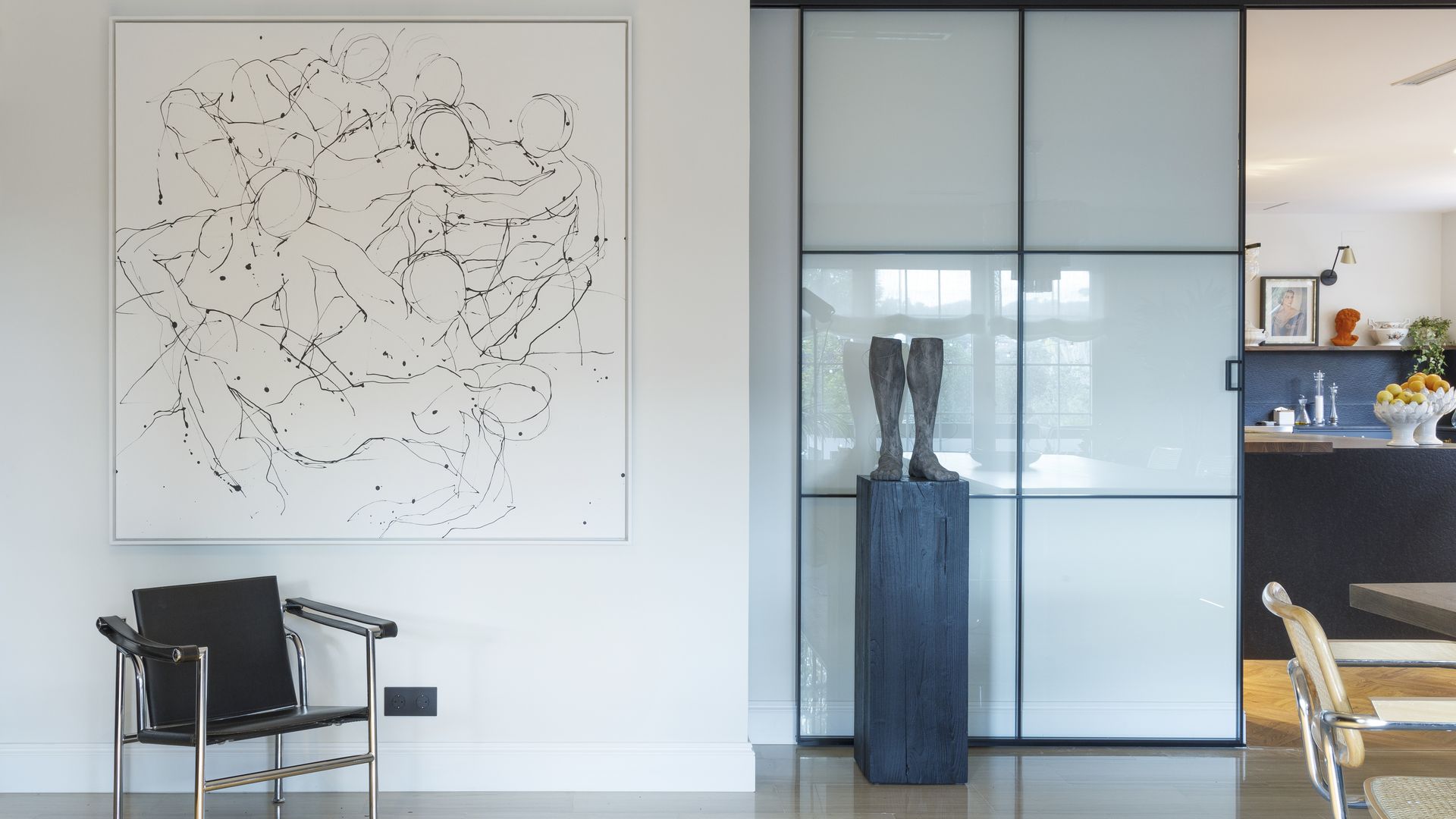 Comedor con silla tipo Wassily, puerta corredera con cuarterones de cristal y perfiles negros, suelo de madera, cuadro en la pared, pedestal con escultura de botas, alfombra