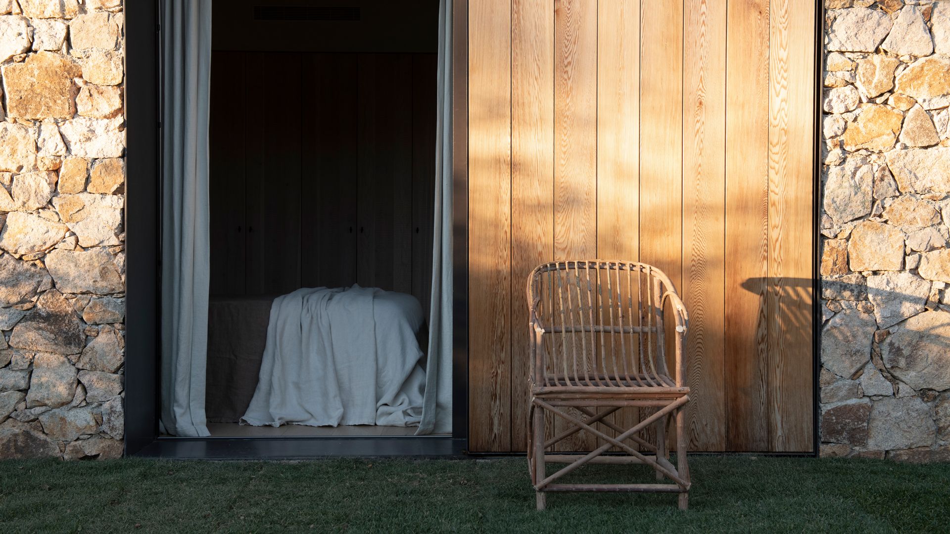 Vista de dormitorio desde el exterior con tabique de lamas de madera, cortinas blancas, sillón de fibras vegetales 