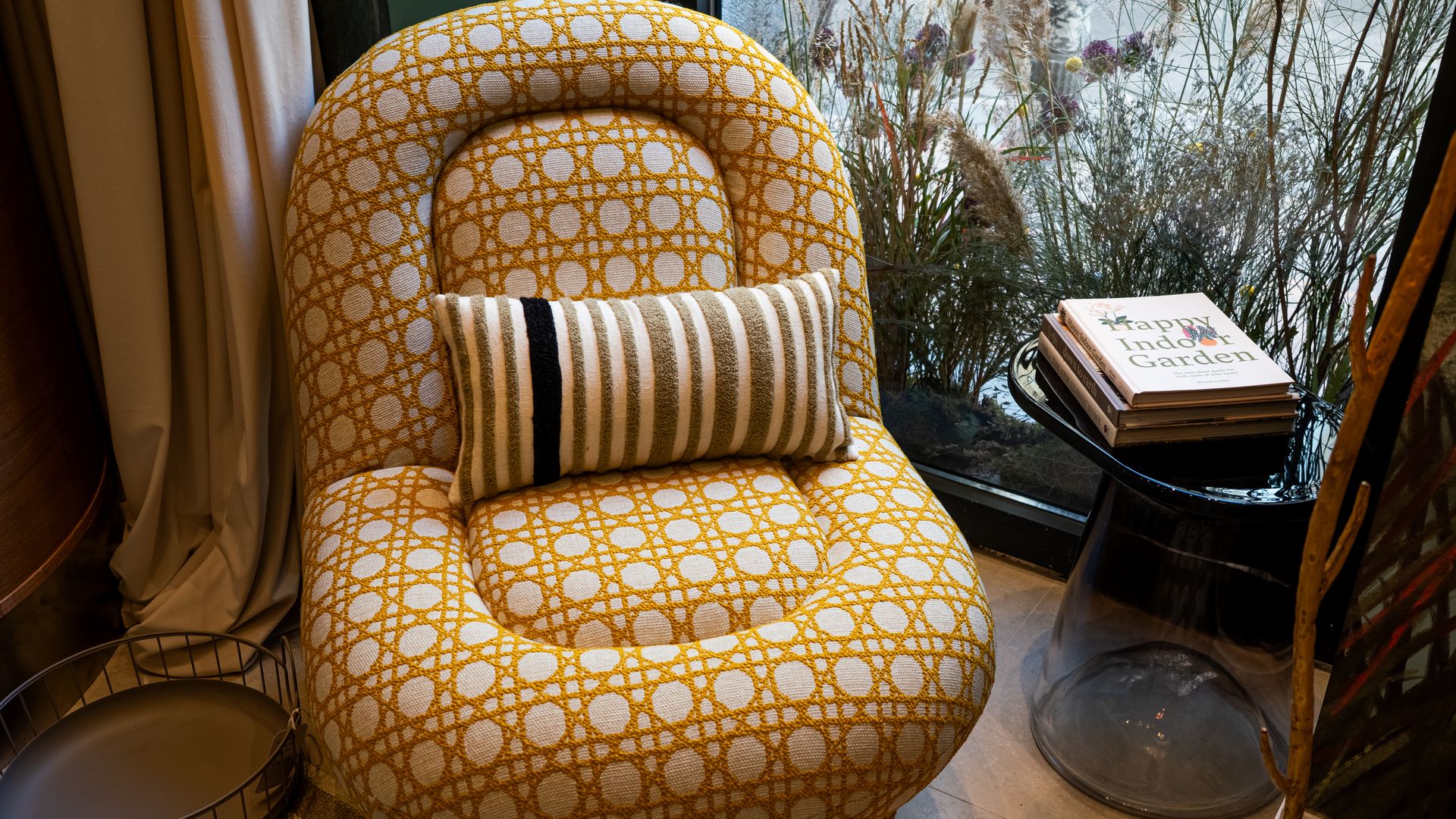 Rincón con de lectura butaca con estampado geométrico e amarillo y blanco, mesa auxiliar negra con libros, ventanal con flores