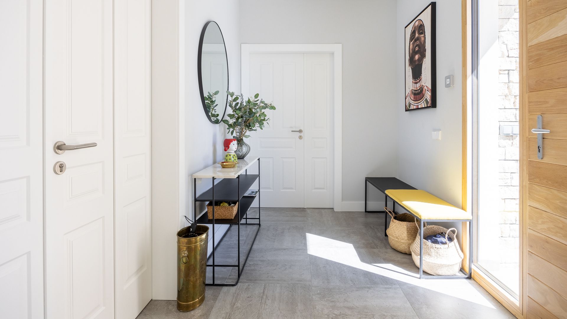 Recibidor con puerta de entrada de madera con tabique de cristal, consola negra, espejo redondo, bancos, cestos bajo el banco, cuadro en la pared, paragüero 