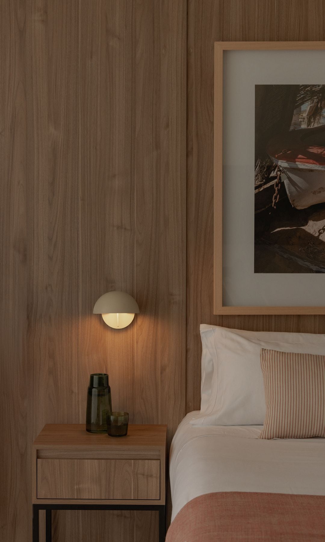 Dormitorio con pared del cabecero de madera, cuadro en la pared, aplique blanco sobre la mesita de noche, mesita de noche de madera, vaso de agua de cristal negro