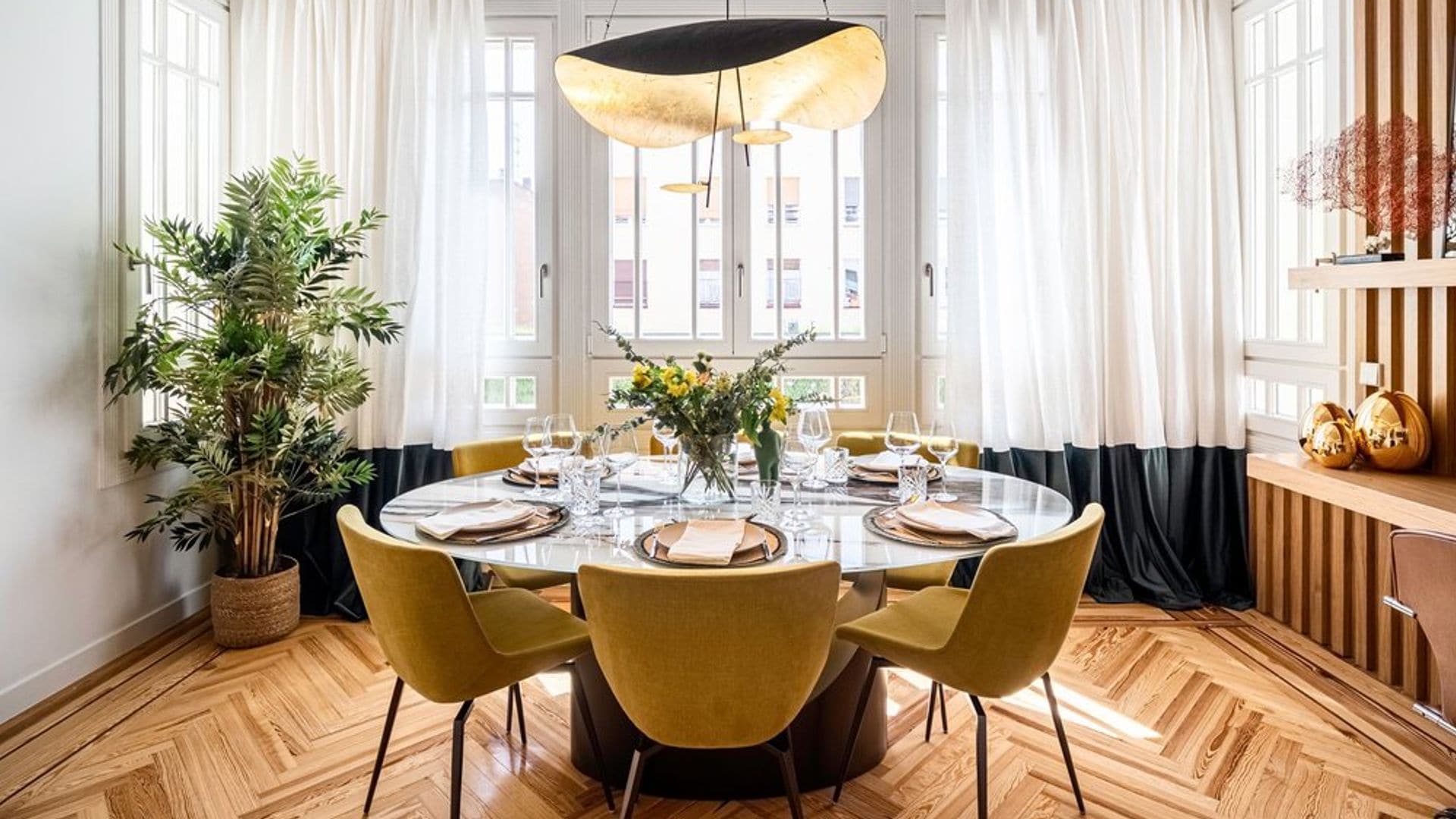 Comedor con suelo de madera en espiga, mesa redonda, lámpara de techo, molduras, cortinas blancas con bajo negro, estantería de madera 