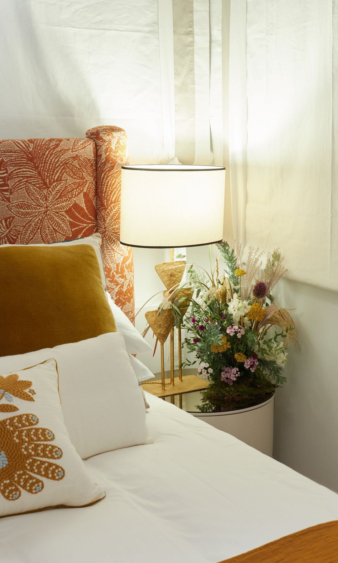 Dormitorio con cabecero tapizado ,mesita de noche, lámpara de sobremesa, arreglo floral, cojines en tonos marrones y blancos 