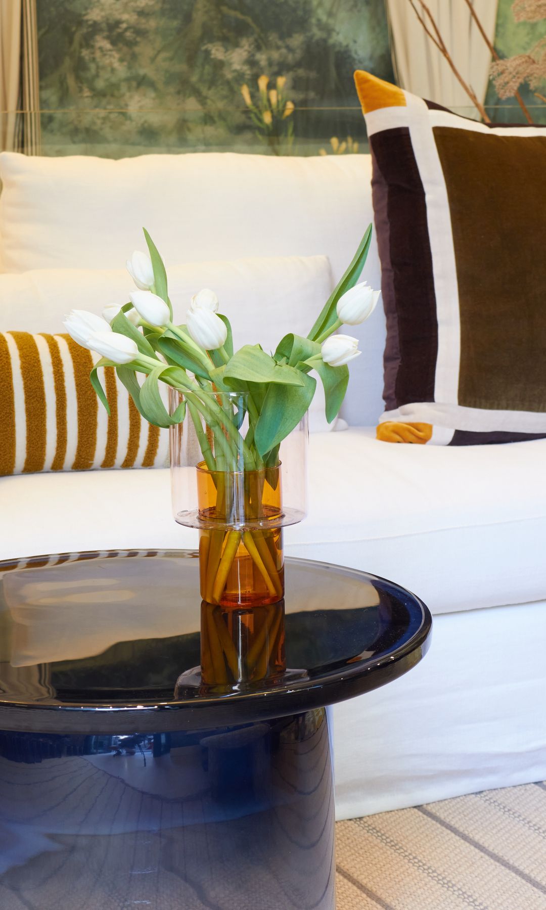 Salón con sofá blanco, mesa auxiliar negra, jarrón de cristal con flores blancas, cojines en tonos marrones 