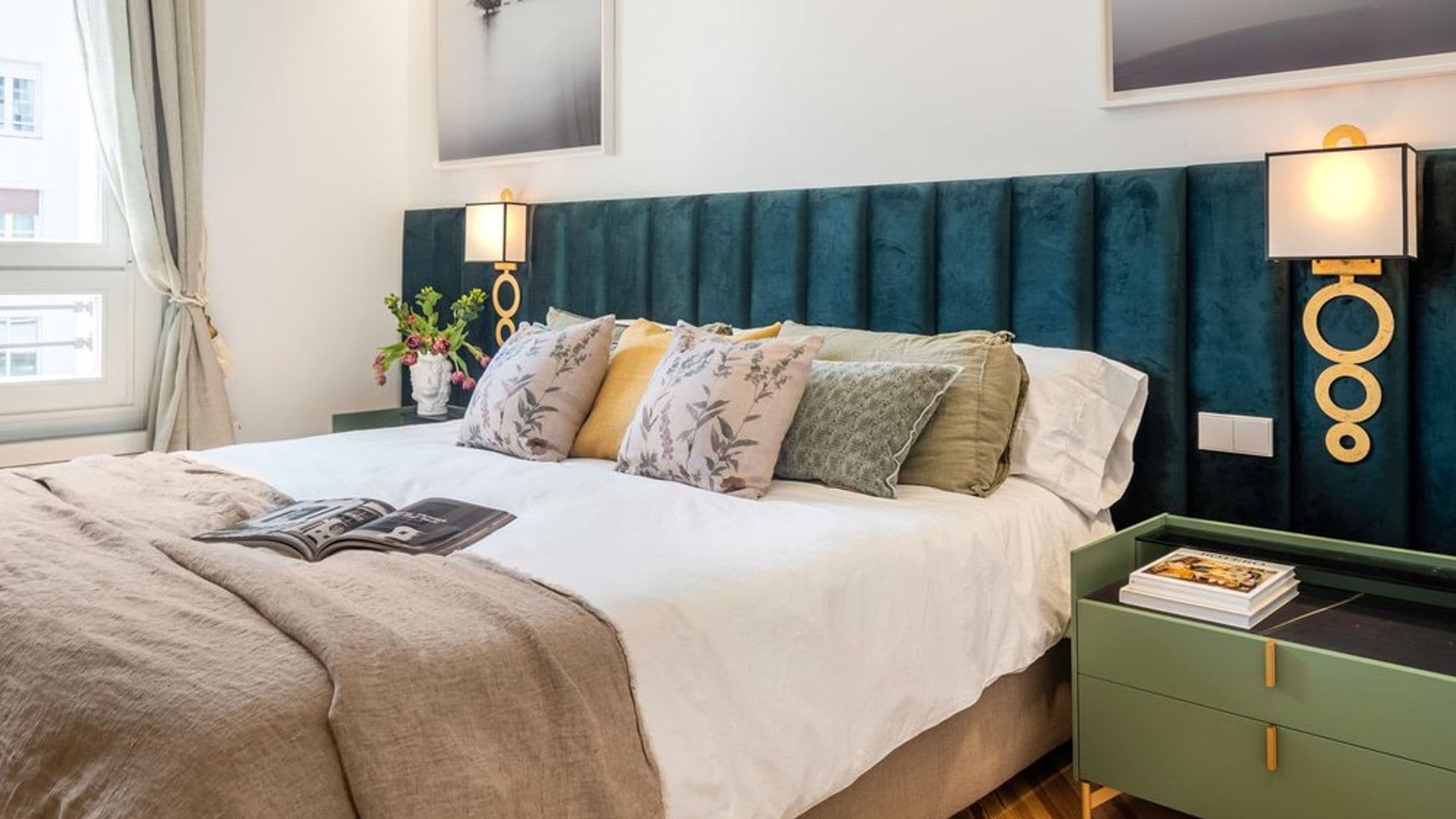 Dormitorio con cabecero tapizado en terciopelo azul oscuro, apliques de pared dorados, mesita de noche verde, cojines sobre la cama, cuadros en la pared del cabecero
