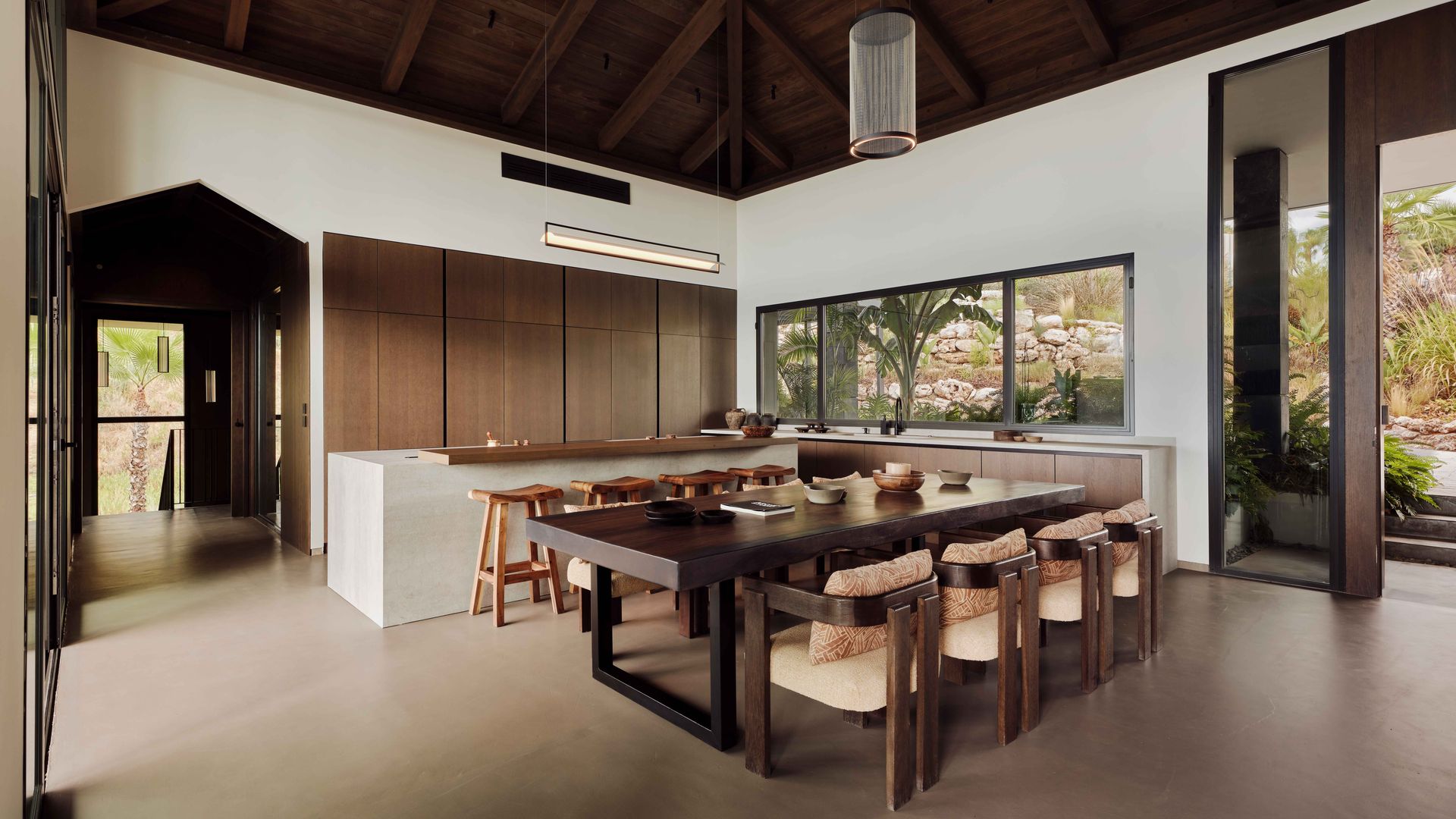Cocina con comedor con gran mesa de madera oscura, con sillas de madera, con respaldo y asiento tapizados, isla de cocina, techo de madera