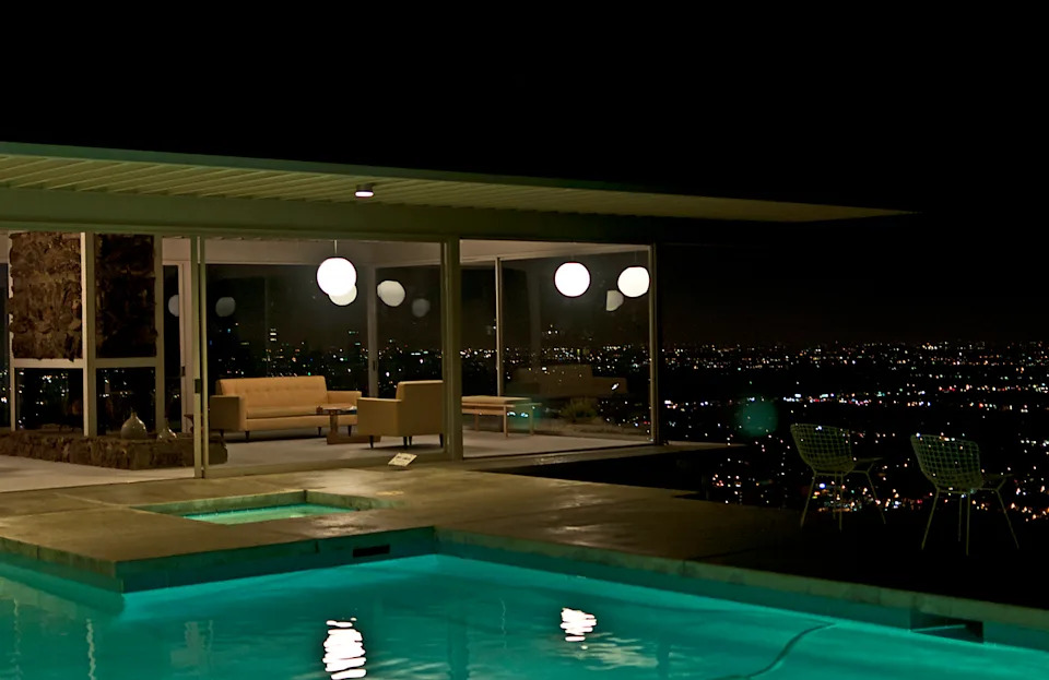 Corner of house with glass walls and flat roof with long eaves perched above nighttime city skyscape.