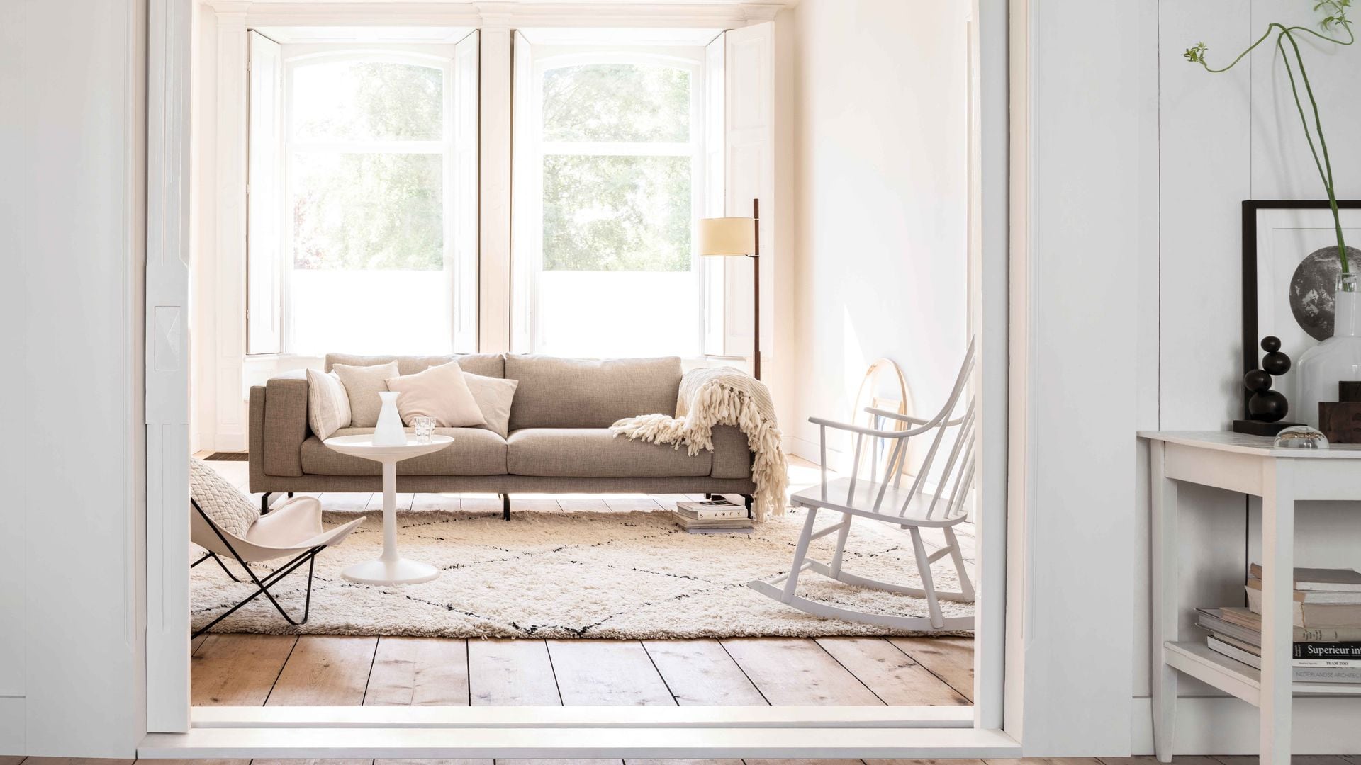 Salón con puerta corredera abierta de color blanco, suelo de madera de lamas anchas, paredes blancas, moldura en el techo, sofá gris, alfombra de pelo