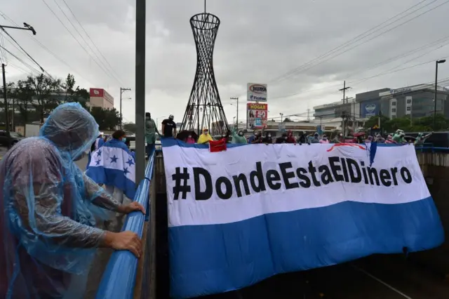 Un grupo de personas sostiene un enorme cartel que dice: ¿Dónde está el dinero?, en el cual tiene los colores de la bandera de Honduras.