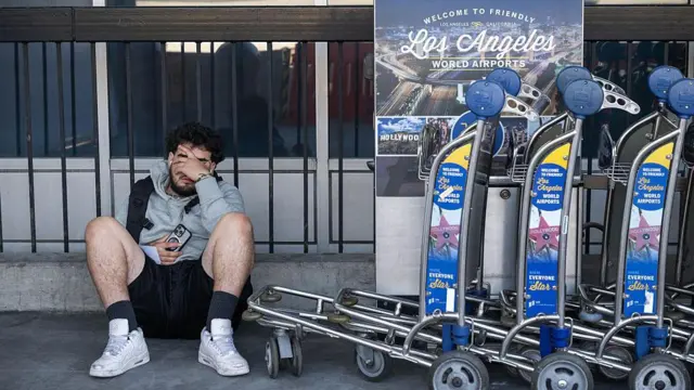 Un hombre sentado en el piso junto a varios carritos para transportar maletas típicas de los aeropuertos. El hombre tiene la mano sobre el rostro con un gesto que denota agotamiento.