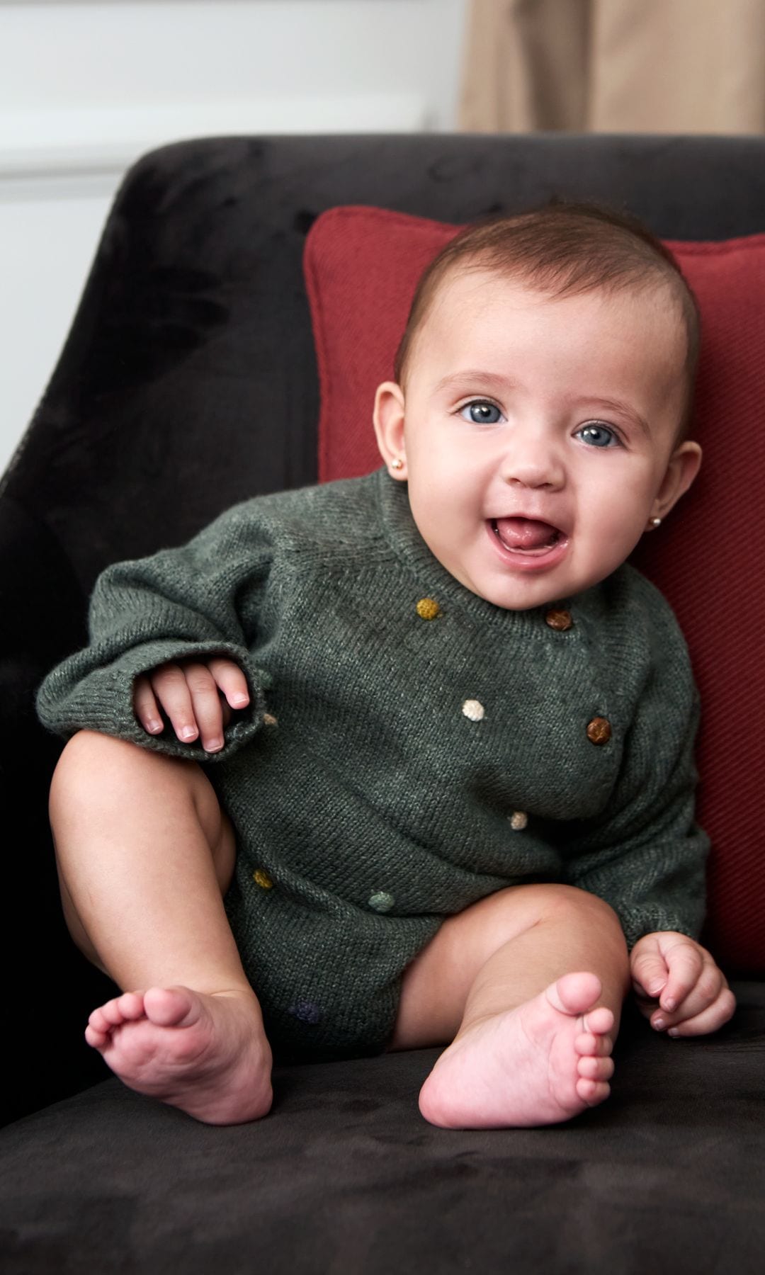 Bebé sonriente en sofá negro con cojín rojo, usando jersey verde