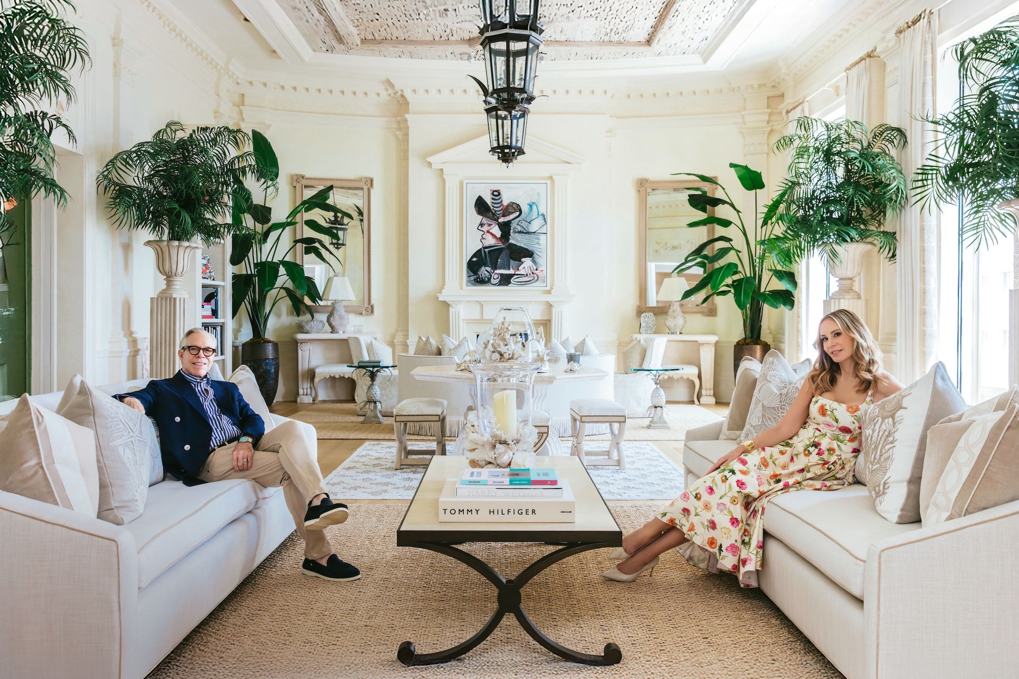 Tommy Hilfiger y su mujer Dee sentados los sofas de su salón blanco con mesa en el centro y chimenea al fondo decorado con plantas en los laterales 
