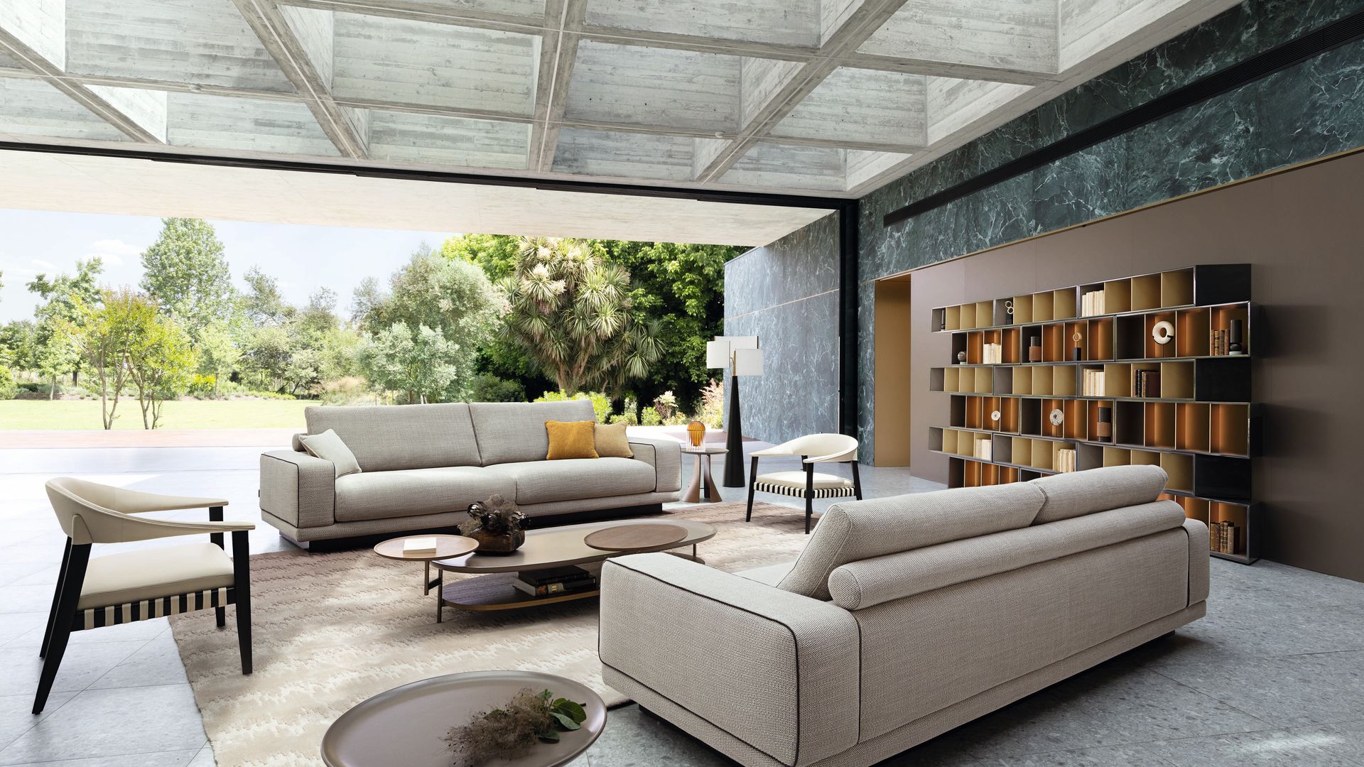 Salón conectado con el jardín, con sofás grises, suelo gris, alfombra, estantería, techo de cemento con aberturas
