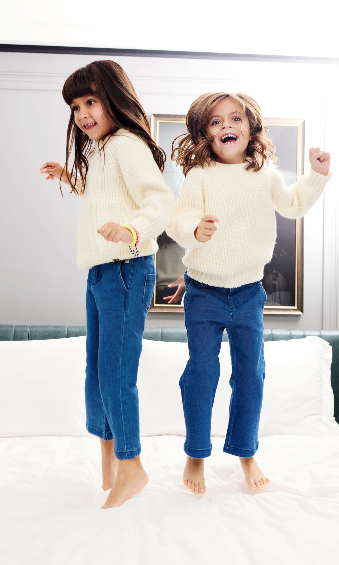 Dos niñas saltan en una cama, ambas con suéteres blancos y jeans azules